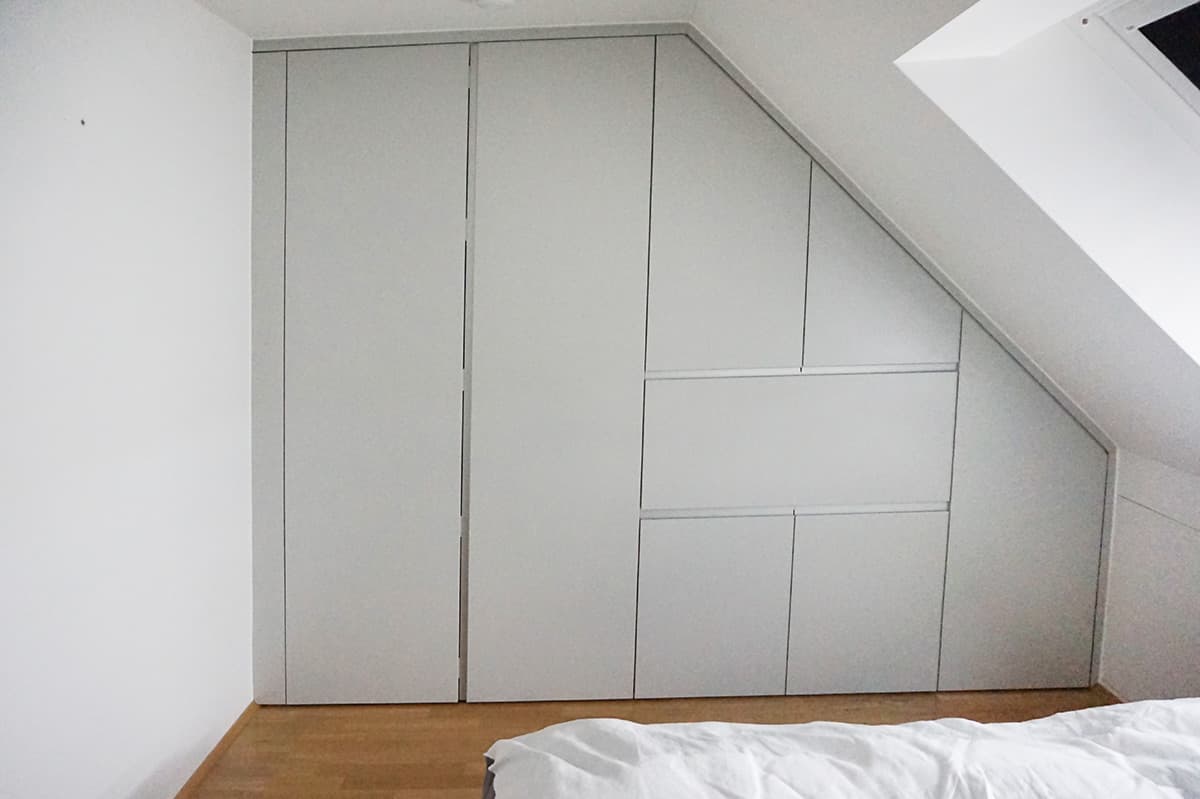 Einbauschrank mit hellen, fugenlos schließenden Fronten, passgenau unter eine Dachschräge gesetzt, daneben ein Dachfenster.